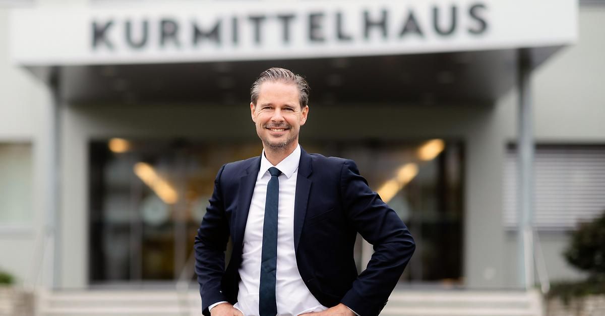 Ein Mann in formeller Kleidung steht vor dem Kurmittelhaus-Gebäude, lächelt und posiert für ein Foto.