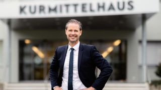 Ein Mann in formeller Kleidung steht vor dem Kurmittelhaus-Gebäude, lächelt und posiert für ein Foto.