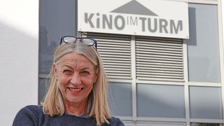 Eine ältere Frau mit blondem Haar lächelt in die Kamera und steht vor einem Gebäude mit dem Schild 'KiNO IM TURM'.