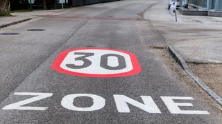 Ein auf den Boden gemalter Verkehrsschild zeigt eine Geschwindigkeitsbegrenzung von 30 km/h. Darunter steht das Wort 'ZONE' in großen weißen Buchstaben.