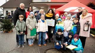 Eine Gruppe von Erwachsenen und Kindern ist draußen versammelt. Die Frau in der Mitte hält einen großen Scheck. Die Kinder tragen Winterkleidung und halten Papiere. Dahinter steht ein Gebäude und ein rotes Zelt.