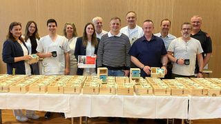 Eine Gruppe von Menschen steht hinter einem Tisch voller Holzkisten, einige halten Gegenstände und andere zeigen Tassen. Sie sind in verschiedenen Outfits gekleidet.