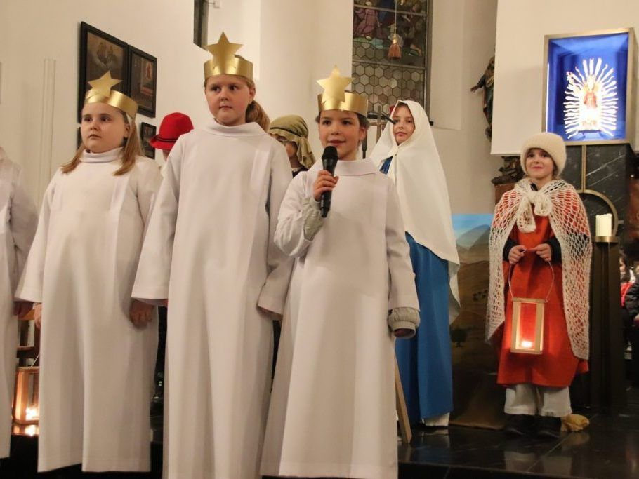 Kinder in weißen Roben mit goldenen Kronen auf dem Kopf stehen auf einer Bühne. Ein Kind hält ein Mikrofon, während ein anderes eine Laterne hält. Die Umgebung scheint eine Kirche zu sein.