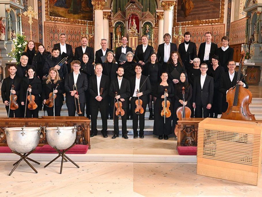 Ein Chor und ein Orchester, in formeller Kleidung, stehen auf der Bühne, halten Instrumente und haben einen hölzernen Hintergrund.