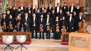 Ein Chor und ein Orchester, in formeller Kleidung, stehen auf der Bühne, halten Instrumente und haben einen hölzernen Hintergrund.