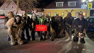Eine Gruppe von Menschen in Kostümen mit Hörnern und Masken steht bei Nacht vor einem Gebäude. Sie halten Stöcke und ein rotes Banner.