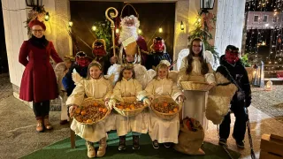 Eine Gruppe von Kindern in Engelskostümen mit Obstkörben, dahinter ein Mann als Weihnachtsmann und zwei Personen in Dämonenmasken.