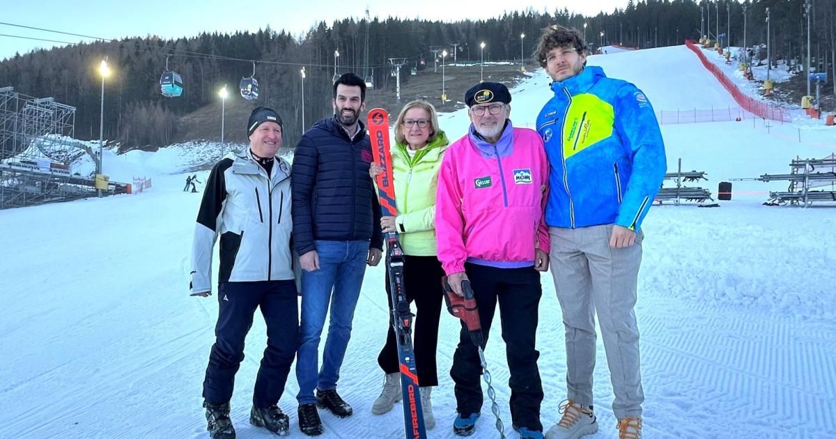 Fünf Personen posieren für ein Foto auf einer verschneiten Piste, mit Skiausrüstung in der Hand. Hinter ihnen sind Bäume und Skilifte zu sehen.