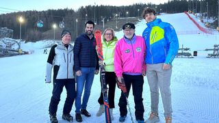 Fünf Personen posieren für ein Foto auf einer verschneiten Piste, mit Skiausrüstung in der Hand. Hinter ihnen sind Bäume und Skilifte zu sehen.