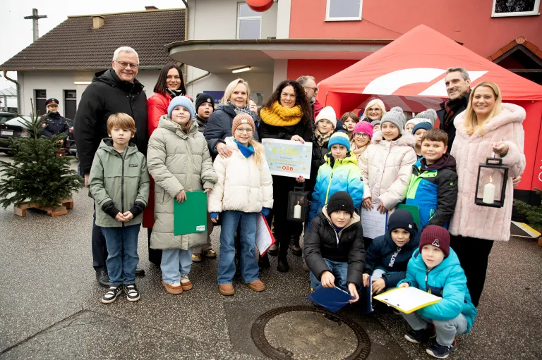 Eine Gruppe von Menschen, darunter Erwachsene und Kinder, posiert für ein Foto im Freien. Einige halten ein Zertifikat und Bücher. Dahinter ist ein rotes Zelt und ein Gebäude mit Schornstein zu sehen.