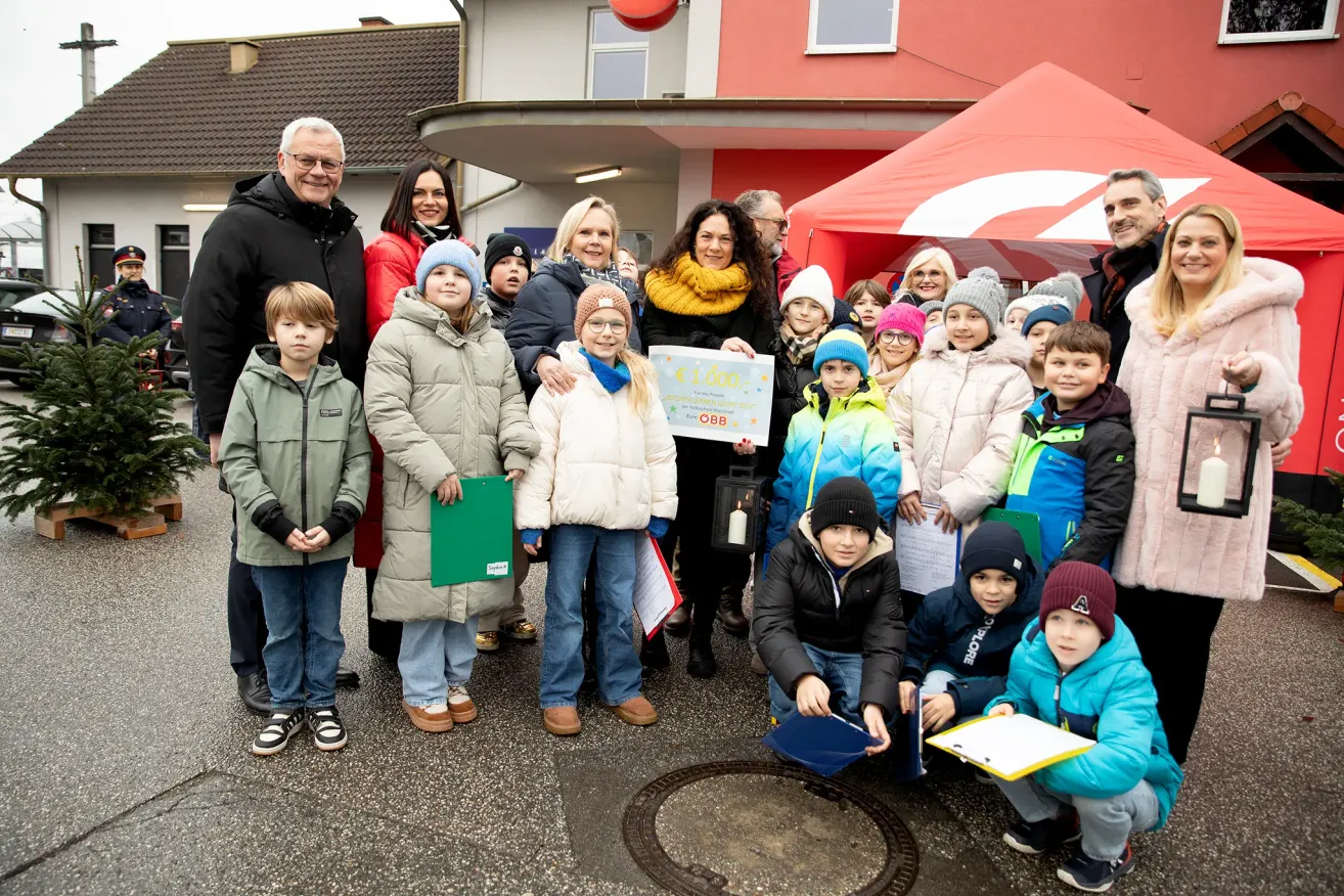 Eine Gruppe von Menschen, darunter Erwachsene und Kinder, posiert für ein Foto im Freien. Einige halten ein Zertifikat und Bücher. Dahinter ist ein rotes Zelt und ein Gebäude mit Schornstein zu sehen.