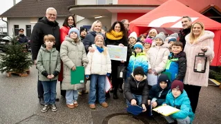 Eine Gruppe von Menschen, darunter Erwachsene und Kinder, posiert für ein Foto im Freien. Einige halten ein Zertifikat und Bücher. Dahinter ist ein rotes Zelt und ein Gebäude mit Schornstein zu sehen.