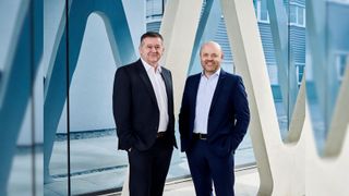 Zwei Männer in Anzügen stehen lächelnd nebeneinander vor einem modernen Gebäude mit blauen und weißen Wänden.