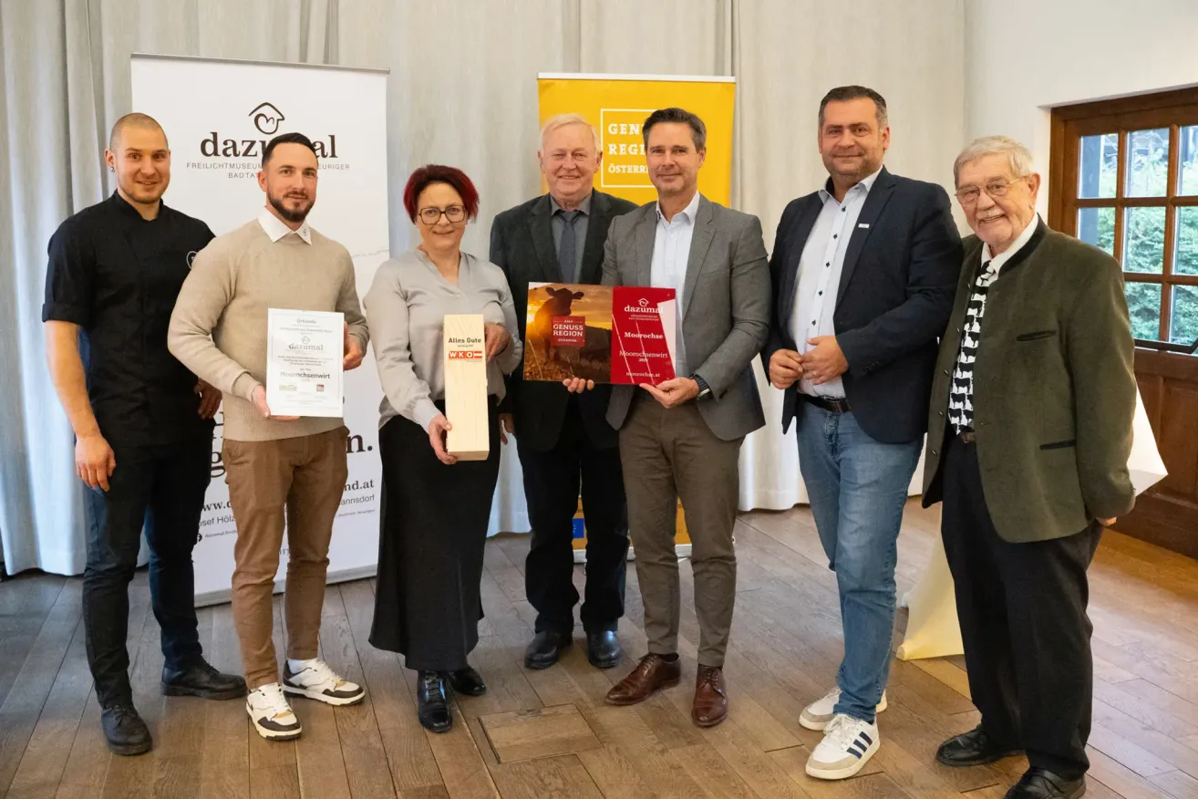 Sechs Personen stehen auf einem Holzboden und lächeln. Drei Männer und drei Frauen, alle halten Urkunden. Dahinter befindet sich ein gelbes Banner mit der Aufschrift 'Genu Region Osterreich'.