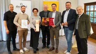 Sechs Personen stehen auf einem Holzboden und lächeln. Drei Männer und drei Frauen, alle halten Urkunden. Dahinter befindet sich ein gelbes Banner mit der Aufschrift 'Genu Region Osterreich'.
