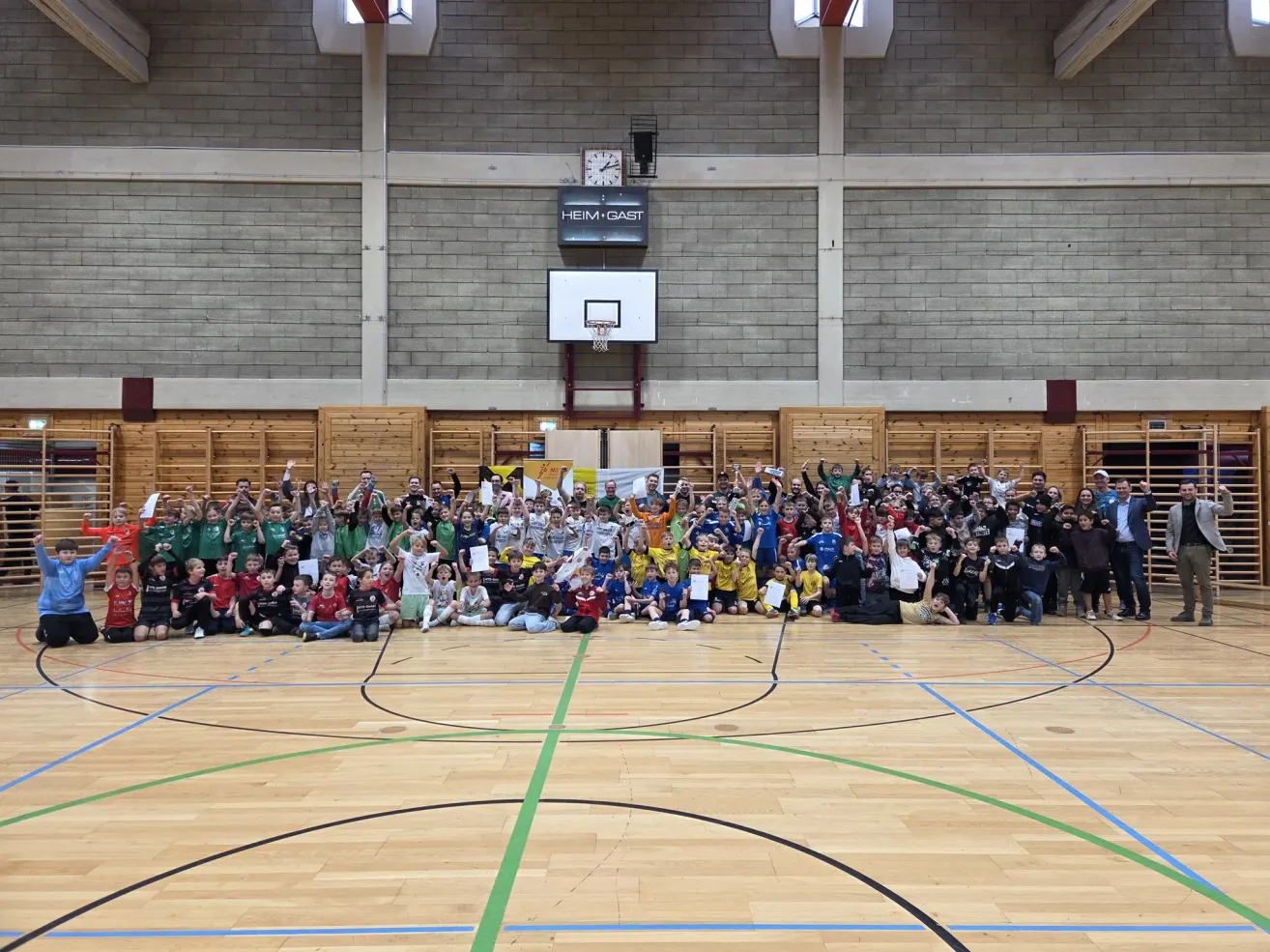 Eine große Gruppe von Kindern in grünen, gelben und roten Trikots versammelt sich in einer Turnhalle mit einem Basketballplatz. Sie halten Urkunden und posieren für ein Foto.