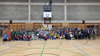 Eine große Gruppe von Kindern in grünen, gelben und roten Trikots versammelt sich in einer Turnhalle mit einem Basketballplatz. Sie halten Urkunden und posieren für ein Foto.