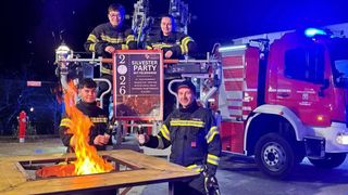 Vier Feuerwehrleute stehen neben einem Feuerwehrauto, zwei halten Flaschen und einer hat ein Schild mit der Aufschrift Silvester Party mit Feuerwehr.