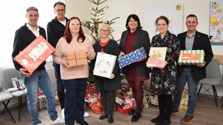 Eine Gruppe von Menschen posiert für ein Foto, während sie Geschenke halten, umgeben von einem Weihnachtsbaum und vielen verpackten Geschenken.