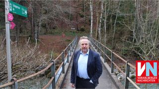 Ein Mann in einem Anzug steht auf einer Holzbrücke über einem Fluss, mit einem Wald auf beiden Seiten.