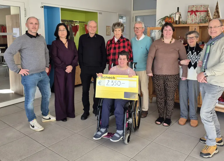 Eine Gruppe von Menschen steht in einem Raum mit einer Person im Rollstuhl, die einen großen gelben Scheck hält. Hinter ihnen befinden sich Regale mit verschiedenen Gegenständen.