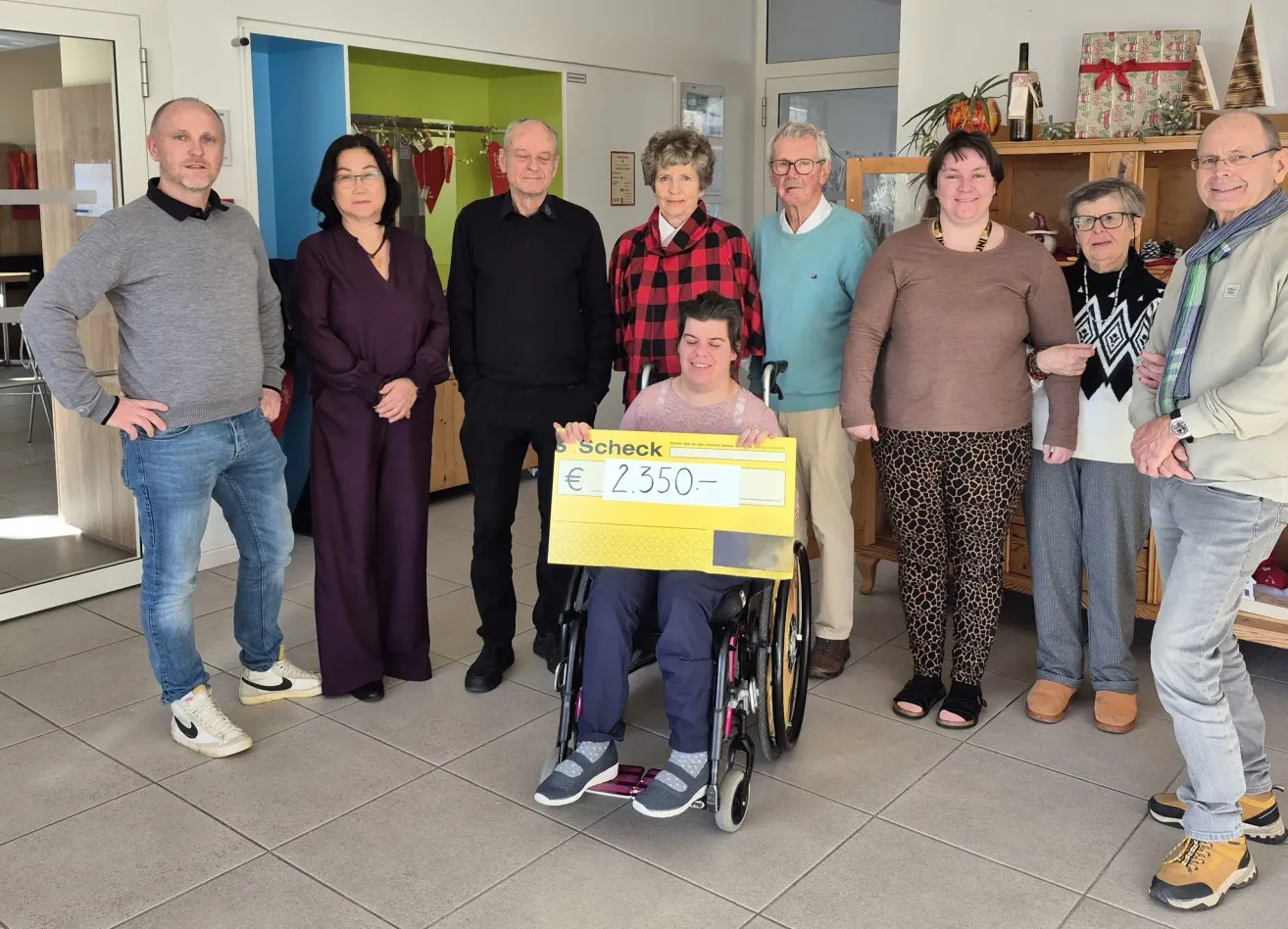 Eine Gruppe von Menschen steht in einem Raum mit einer Person im Rollstuhl, die einen großen gelben Scheck hält. Hinter ihnen befinden sich Regale mit verschiedenen Gegenständen.