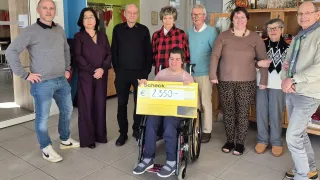 Eine Gruppe von Menschen steht in einem Raum mit einer Person im Rollstuhl, die einen großen gelben Scheck hält. Hinter ihnen befinden sich Regale mit verschiedenen Gegenständen.