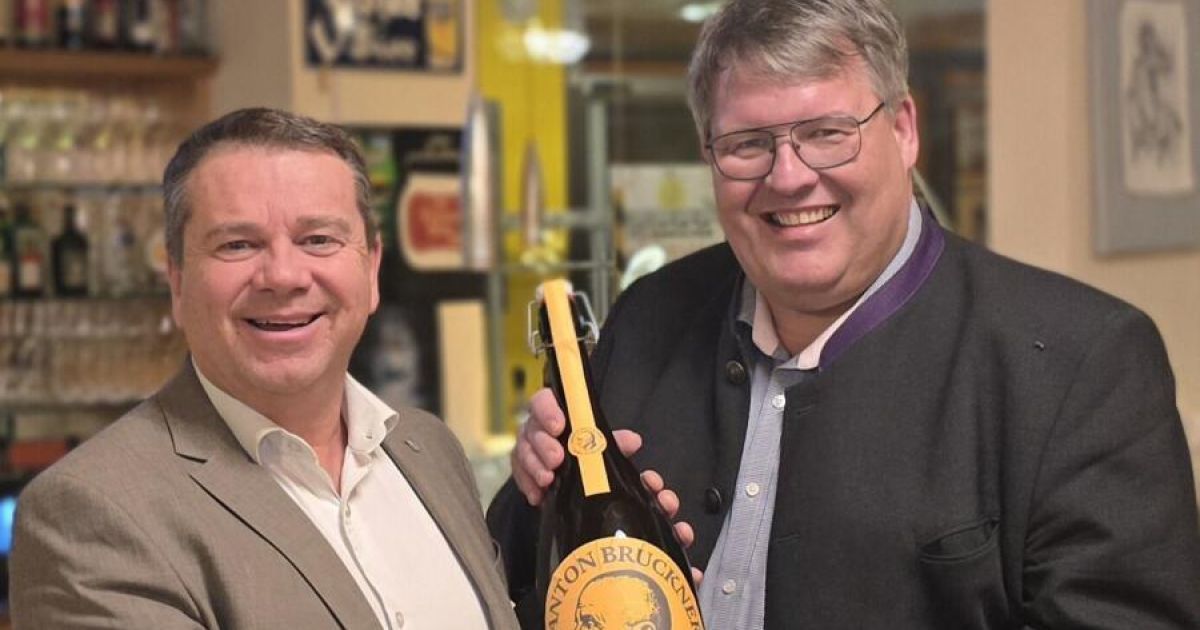 Zwei Männer lächeln, einer hält eine Flasche Nanton Bruckwein, beide tragen eine Brille, einer in einem Anzug und der andere in einer Jacke.