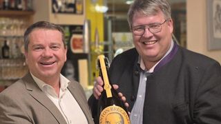 Zwei Männer lächeln, einer hält eine Flasche Nanton Bruckwein, beide tragen eine Brille, einer in einem Anzug und der andere in einer Jacke.