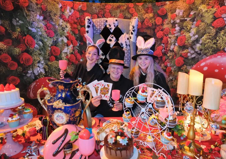 Drei Personen in Alice im Wunderland-Kostümen sitzen an einem Tisch. Ein Mann hält einen Becher und zwei Frauen lächeln. Der Tisch ist mit Kuchen, Cupcakes, einer Vase und einem Weinglas geschmückt. Ein floraler Hintergrund und eine kleine Laterne sind im Hintergrund zu sehen.