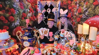 Drei Personen in Alice im Wunderland-Kostümen sitzen an einem Tisch. Ein Mann hält einen Becher und zwei Frauen lächeln. Der Tisch ist mit Kuchen, Cupcakes, einer Vase und einem Weinglas geschmückt. Ein floraler Hintergrund und eine kleine Laterne sind im Hintergrund zu sehen.
