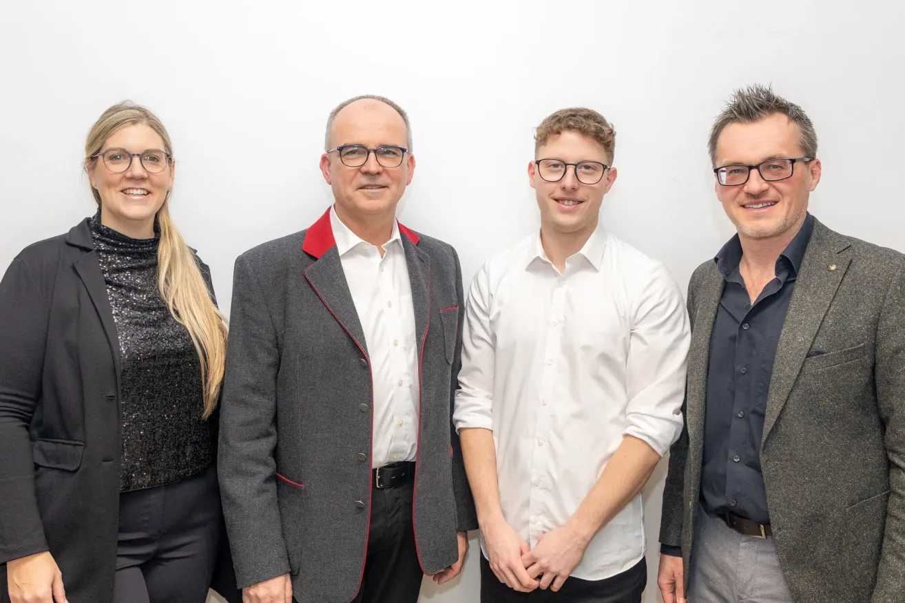 Vier Personen posieren für ein Foto. Sie tragen alle formelle Kleidung. Der Mann links hat einen grauen Mantel mit rotem Besatz. Der Mann neben ihm trägt ein weißes Hemd. Die beiden Männer lächeln.