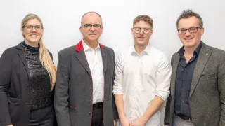 Vier Personen posieren für ein Foto. Sie tragen alle formelle Kleidung. Der Mann links hat einen grauen Mantel mit rotem Besatz. Der Mann neben ihm trägt ein weißes Hemd. Die beiden Männer lächeln.