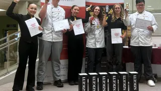 Eine Gruppe von Studenten einer Kochschule steht vor einem Banner und hält Trophäen und Urkunden in den Händen. Sie tragen alle Kochuniformen.