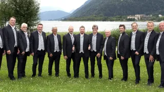 Sieben Männer in Anzügen und Westen posieren für ein Foto auf einem Rasenfeld mit einem See und Bergen im Hintergrund.