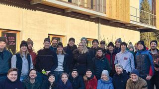 Eine Gruppe von Menschen steht vor einem Gebäude und lächelt für ein Foto. Das Gebäude hat Glasfenster und eine Holzstruktur. Sie tragen Winterkleidung.
