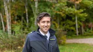 Ein Mann in einer Uniformjacke steht lächelnd in einem Grasbereich mit herbstlichen Bäumen im Hintergrund.
