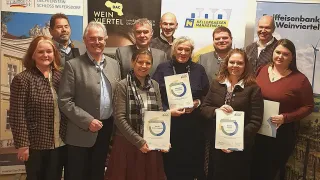 Eine Gruppe von Menschen posiert mit Urkunden vor einem Banner mit einem Logo, einschließlich Namen wie DAC, Kellergassen Management und anderen, wobei einige lächeln und in die Kamera schauen.