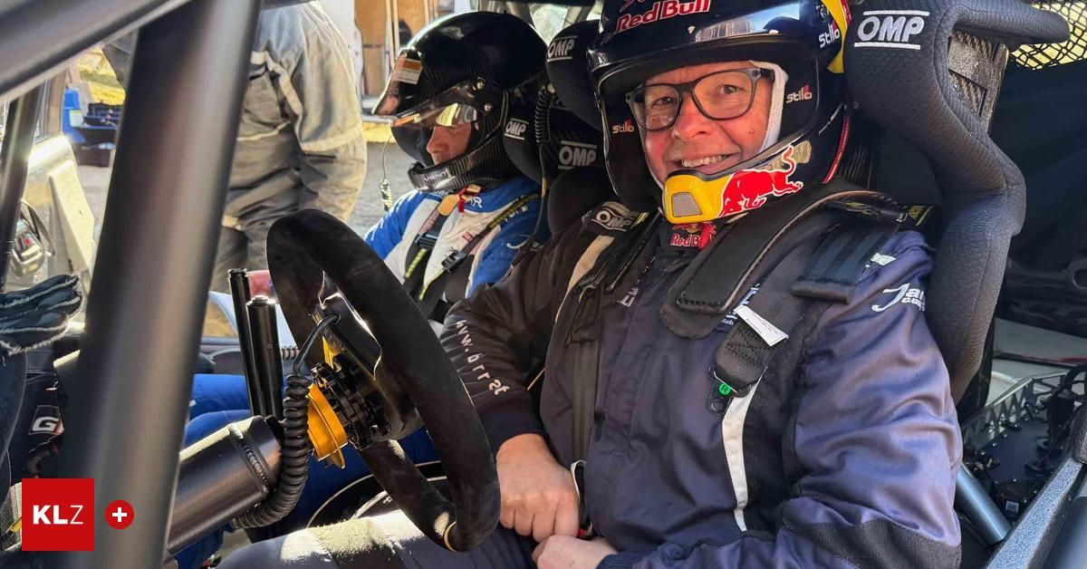 Ein Mann mit einem Red Bull-Helm und einer Brille sitzt in einem Rennwagen. Neben ihm sitzt eine weitere Person, die ebenfalls einen Helm trägt. Das Lenkrad des Autos ist im Vordergrund zu sehen.