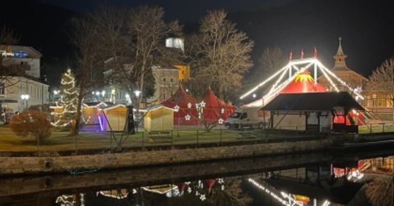 Eine nächtliche Szene zeigt ein Jahrmarkt-Setup am Fluss mit leuchtenden roten Zelten, festlichen Lichtern und einem großen Riesenrad. Bäume säumen das Gebiet, und Gebäude sind in der Ferne sichtbar.