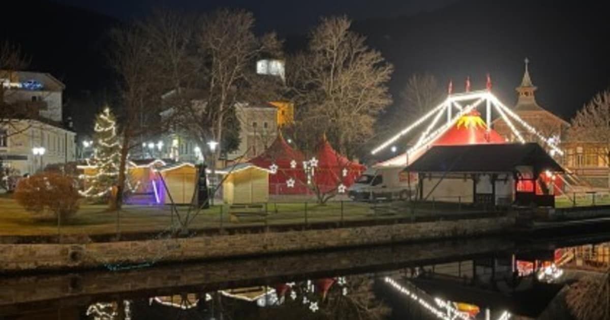 Eine nächtliche Szene zeigt ein Jahrmarkt-Setup am Fluss mit leuchtenden roten Zelten, festlichen Lichtern und einem großen Riesenrad. Bäume säumen das Gebiet, und Gebäude sind in der Ferne sichtbar.