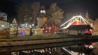 Eine nächtliche Szene zeigt ein Jahrmarkt-Setup am Fluss mit leuchtenden roten Zelten, festlichen Lichtern und einem großen Riesenrad. Bäume säumen das Gebiet, und Gebäude sind in der Ferne sichtbar.