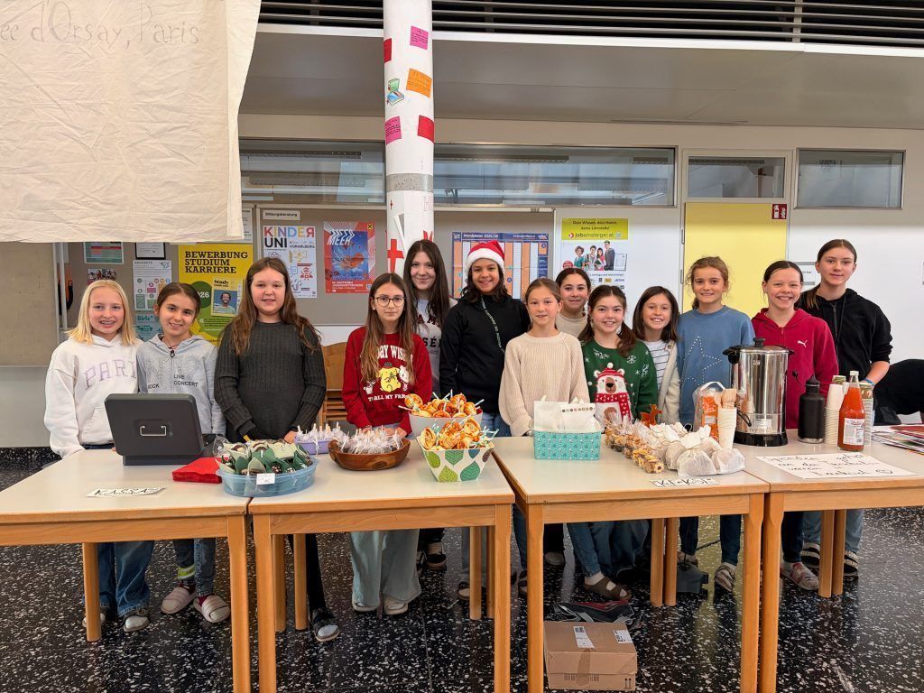 Eine Gruppe junger Mädchen posiert mit Tabletts voller Essen bei einer Schulveranstaltung, trägt festliche Kleidung und lächelt in die Kamera.