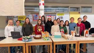 Eine Gruppe junger Mädchen posiert mit Tabletts voller Essen bei einer Schulveranstaltung, trägt festliche Kleidung und lächelt in die Kamera.