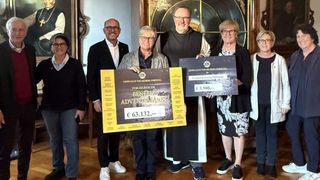 Eine Gruppe von Menschen steht drinnen, lächelt und posiert für ein Foto. Ein Mann hält ein gelbes Banner mit Text, während eine Frau ein Zertifikat in der Hand hat. Hinter ihnen stehen Holzschränke und gerahmte Porträts.