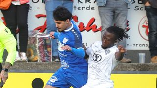 Zwei Fußballspieler prallen während eines Spiels zusammen. Ein Spieler trägt ein blaues Trikot, der andere ein weißes. Im Hintergrund sehen Sie verschwommene Zuschauer und eine Wand mit roten und weißen Graffiti.