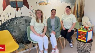 Drei Frauen sitzen auf Stühlen vor einer bunten Wandmalerei. Die Frau auf der linken Seite trägt weiße Hosen und Turnschuhe. Die Frau in der Mitte trägt ein graues Oberteil und eine grüne Strickjacke. Die Frau auf der rechten Seite trägt ein weißes Oberteil und schwarze Hose.