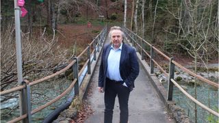 Ein Mann steht auf einer Brücke über einem Fluss, trägt einen blauen Blazer und schwarze Hosen. Die Brücke ist von Metallgeländern umgeben und vor einem Hintergrund von Bäumen.