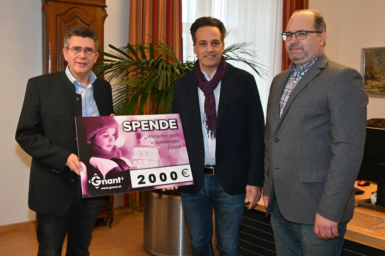 Drei Männer stehen nebeneinander. Der Mann auf der linken Seite hält ein Poster mit einem Mädchen und den Worten 'Spende 2000€'. Dahinter steht eine Pflanze in einem Metalltopf.