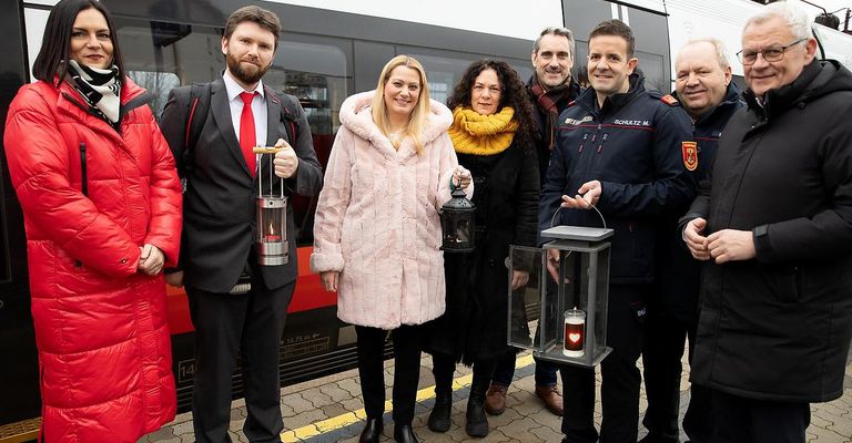 Fünf Personen stehen lächelnd auf einem Bahnsteig. Sie halten Laternen mit Kerzen darin. Ein Zug ist hinter ihnen.